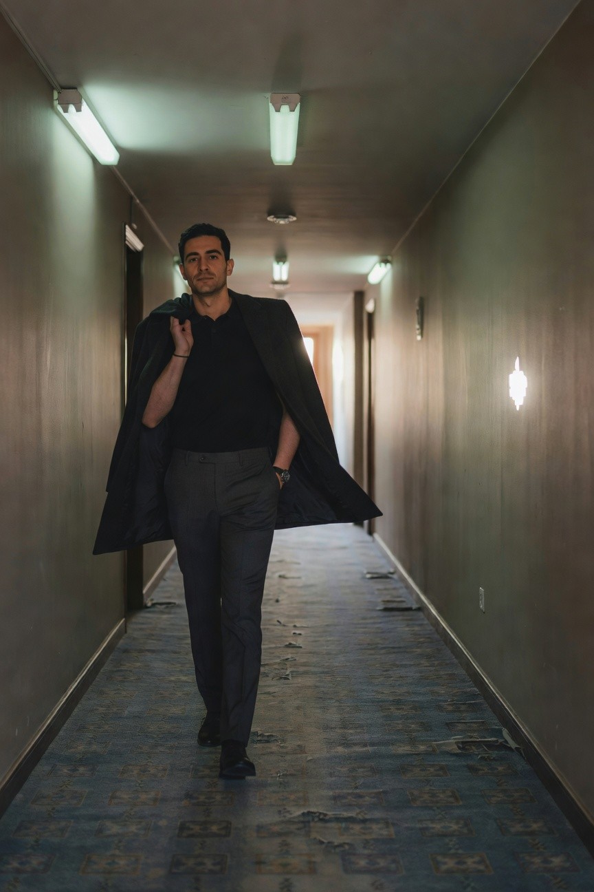 Man walking down hotel hallway in black polo shirt, charcoal grey trousers, black overcoat draped over one shoulder, black dress shoes, and wristwatch, smart casual style