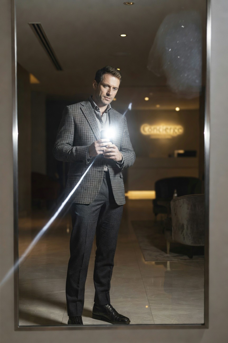 Man in tailored checkered grey blazer, open white shirt, charcoal grey trousers, and black dress shoes reflected in hotel lobby mirror while holding phone with flash lit