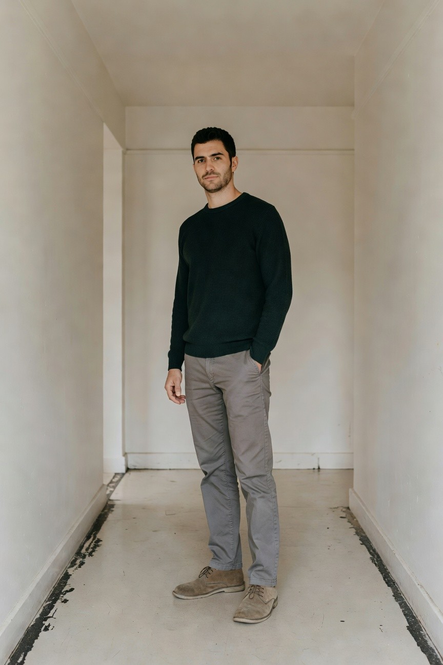 Man in dark green ribbed crewneck sweater, mid-grey slim pants, and tan suede chukka boots standing casually with hands in pockets in a plain hallway