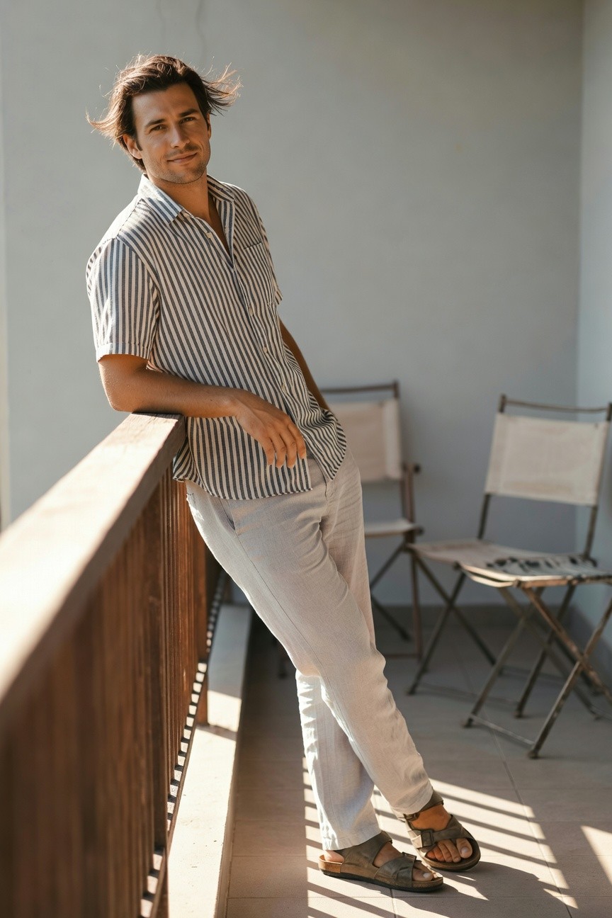 Man in short-sleeve light blue and white striped linen shirt, light grey linen pants, and brown leather sandals, leaning casually on wooden railing