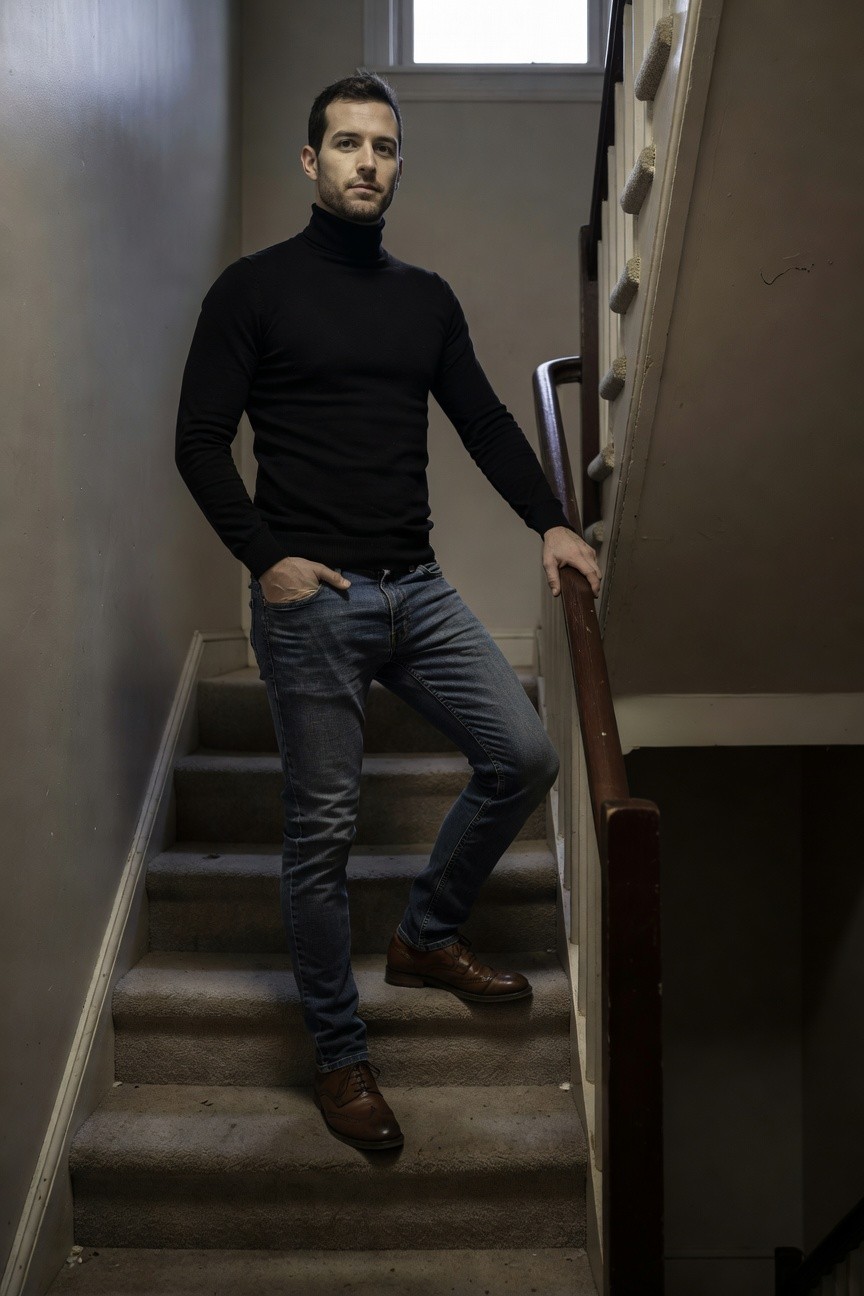 A man stands casually on a carpeted staircase wearing a fitted black turtleneck sweater, dark blue straight-leg jeans, and brown leather dress shoes, one hand in pocket and the other on the wooden railing