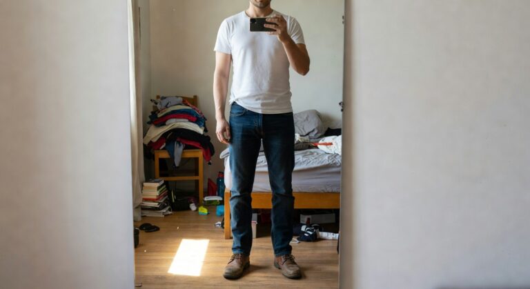 Man in white short-sleeve t-shirt, dark blue straight-leg jeans, and tan leather boots standing in a doorway, casual everyday style
