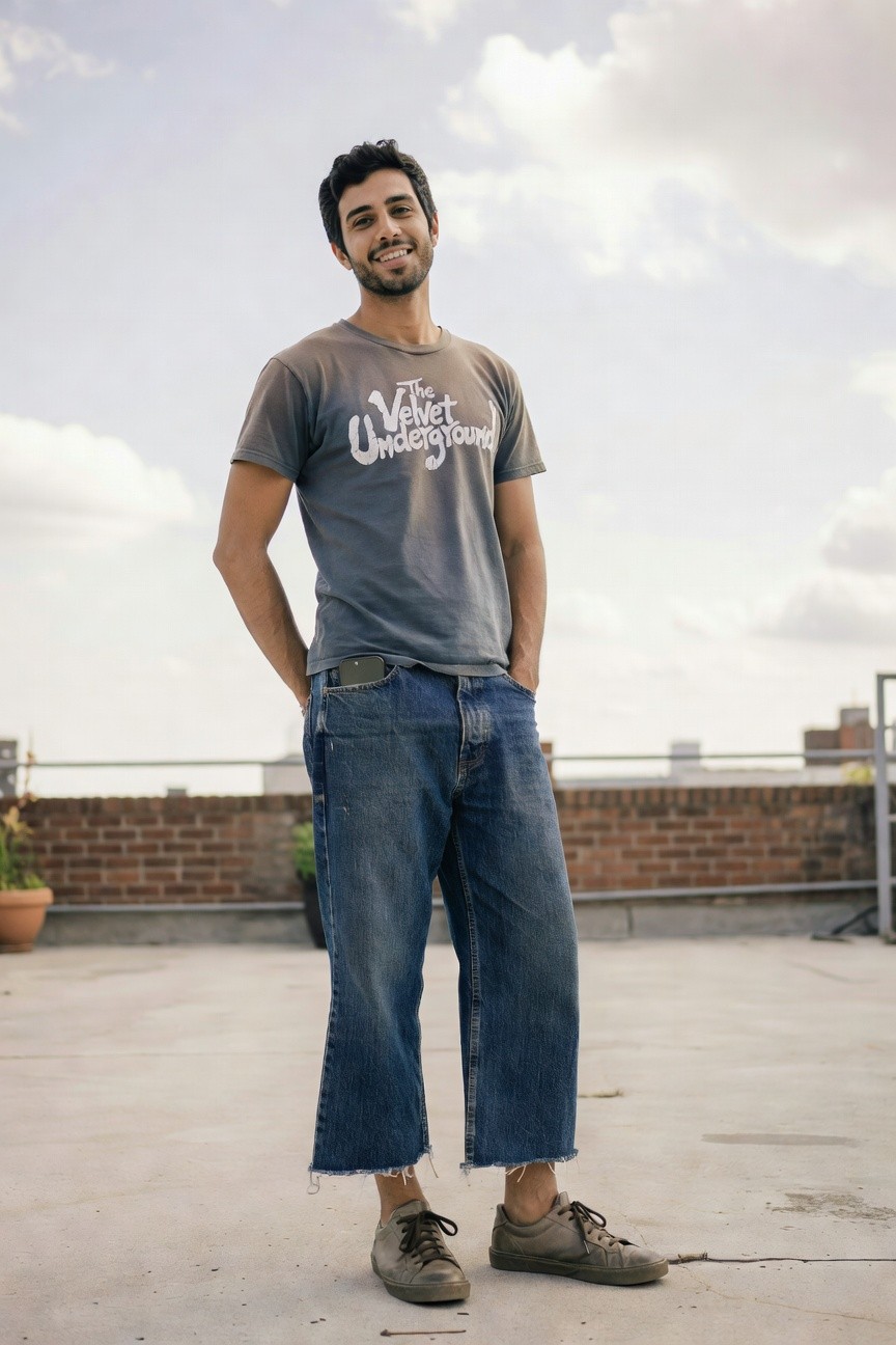 Man in relaxed pose with hands in pockets, wearing a gray short-sleeve Velvet Underground graphic t-shirt, baggy blue cropped jeans with frayed hems, and brown suede sneakers on a concrete surface