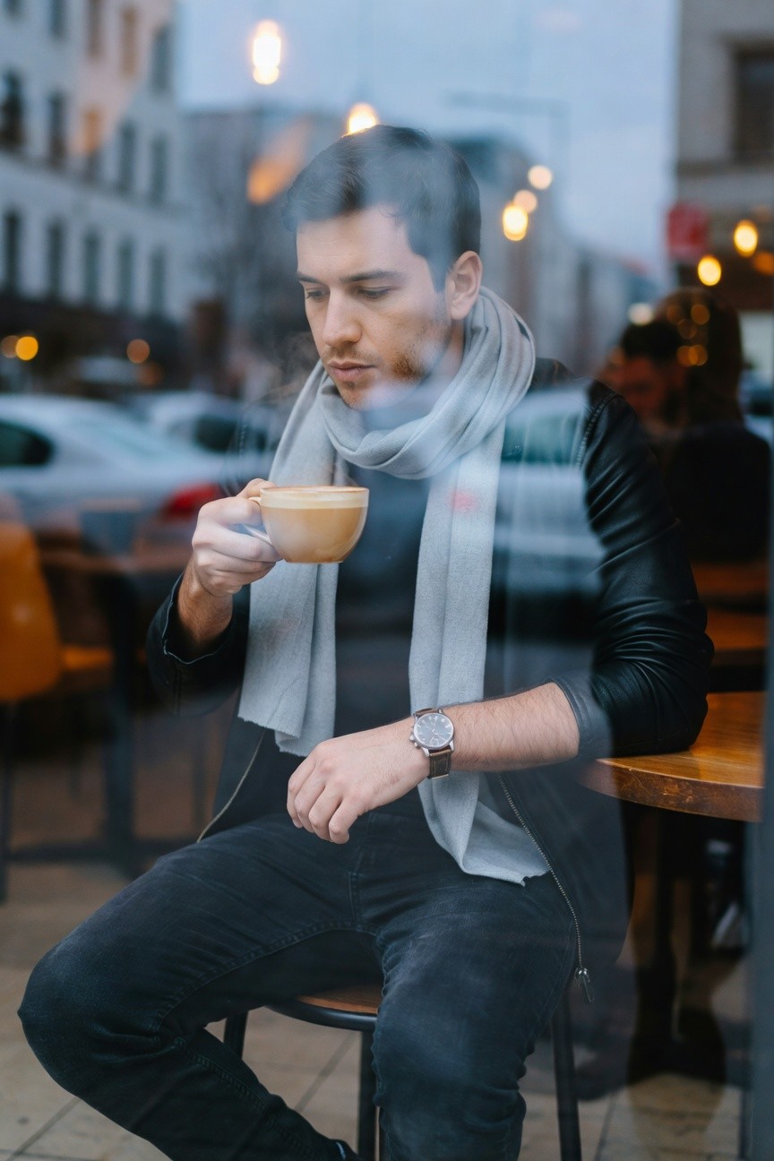 Man in slim black jeans, black leather jacket, beige sweater, and loose gray scarf, sitting at a cafe table holding a coffee cup, viewed through a window
