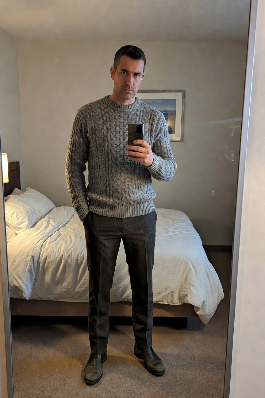 Man in gray cable knit sweater, charcoal wool trousers, and dark Chelsea boots standing in front of hotel mirror selfie, hands in pockets