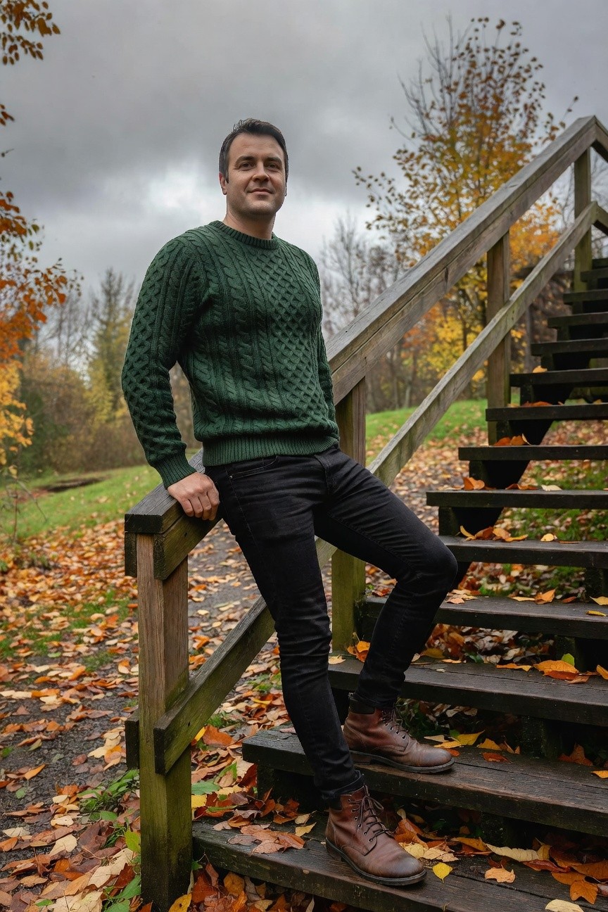 Man leaning on wooden railing wearing green cable knit sweater, black slim jeans, and brown leather boots