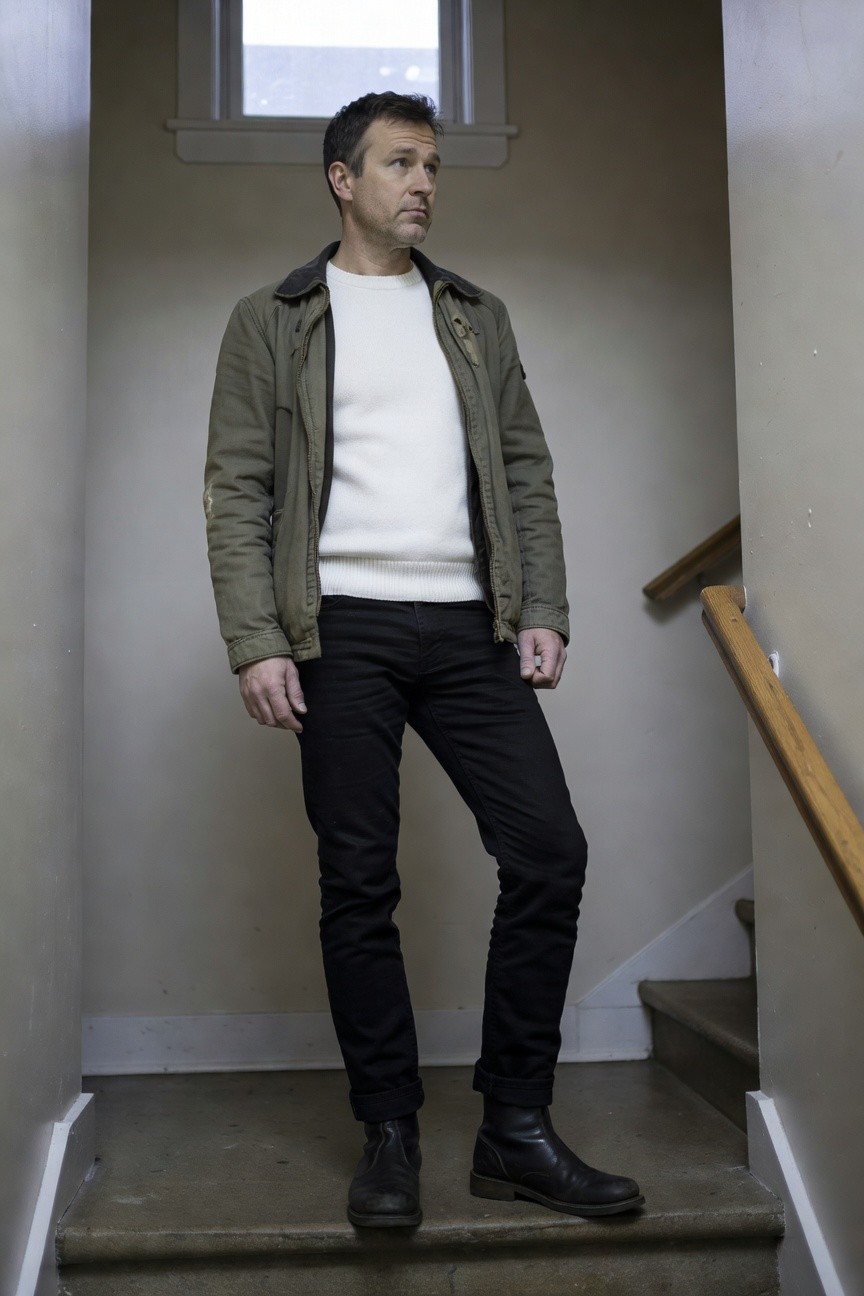 Man standing on indoor stairs in olive green field jacket with shearling collar over white crewneck sweater, black slim pants cuffed at ankles, and black leather Chelsea boots, looking thoughtful