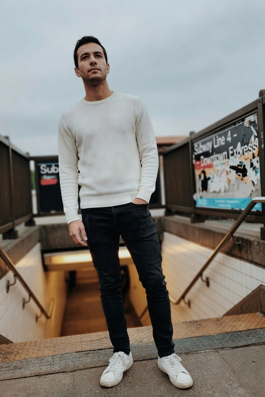 Man in cream crewneck sweater, dark slim-fit jeans, and white sneakers, hands in pockets, standing on stairs