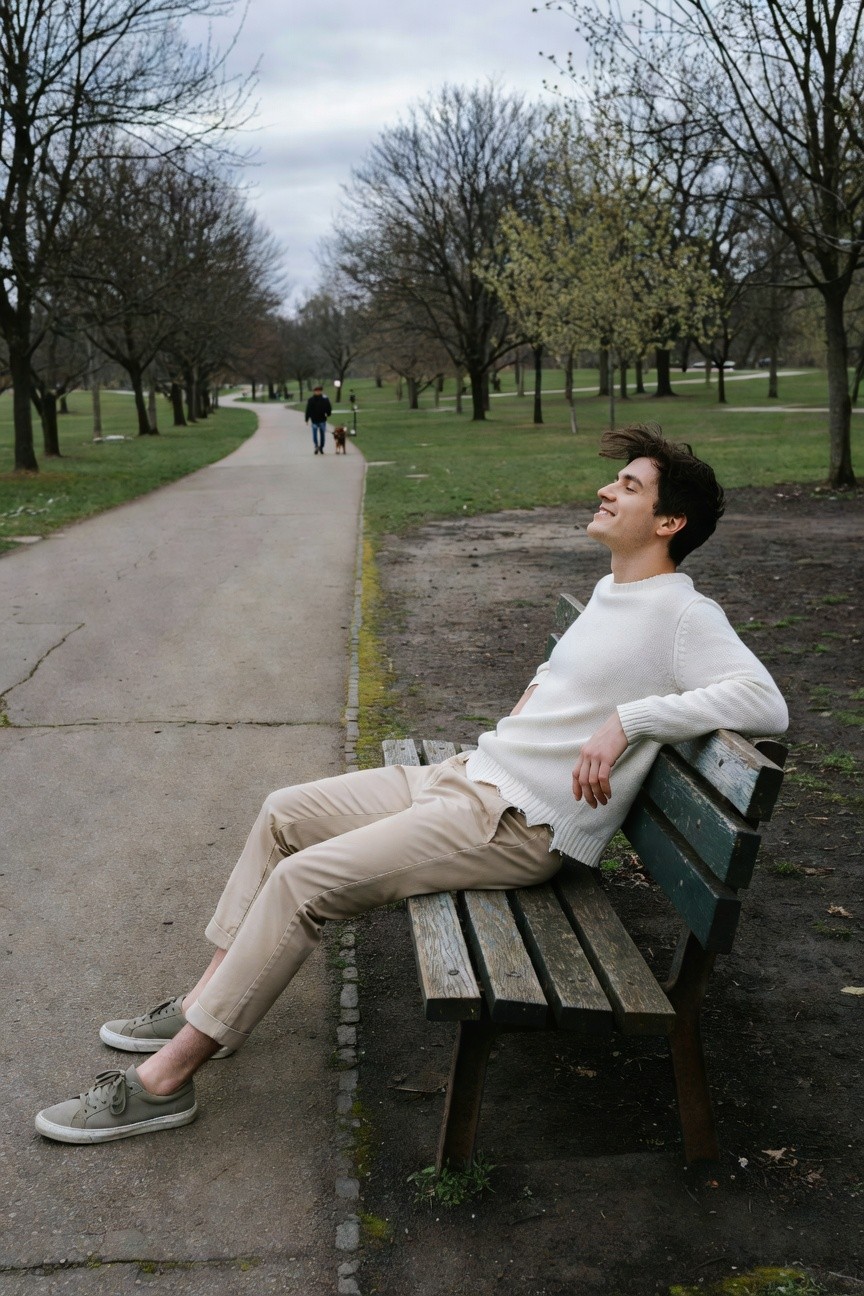 Young man lounging sideways on a wooden park bench wearing a white knit crewneck sweater, slim beige khaki pants cropped at the ankles, and gray sneakers with white soles, smiling with eyes closed against an outdoor path backdrop