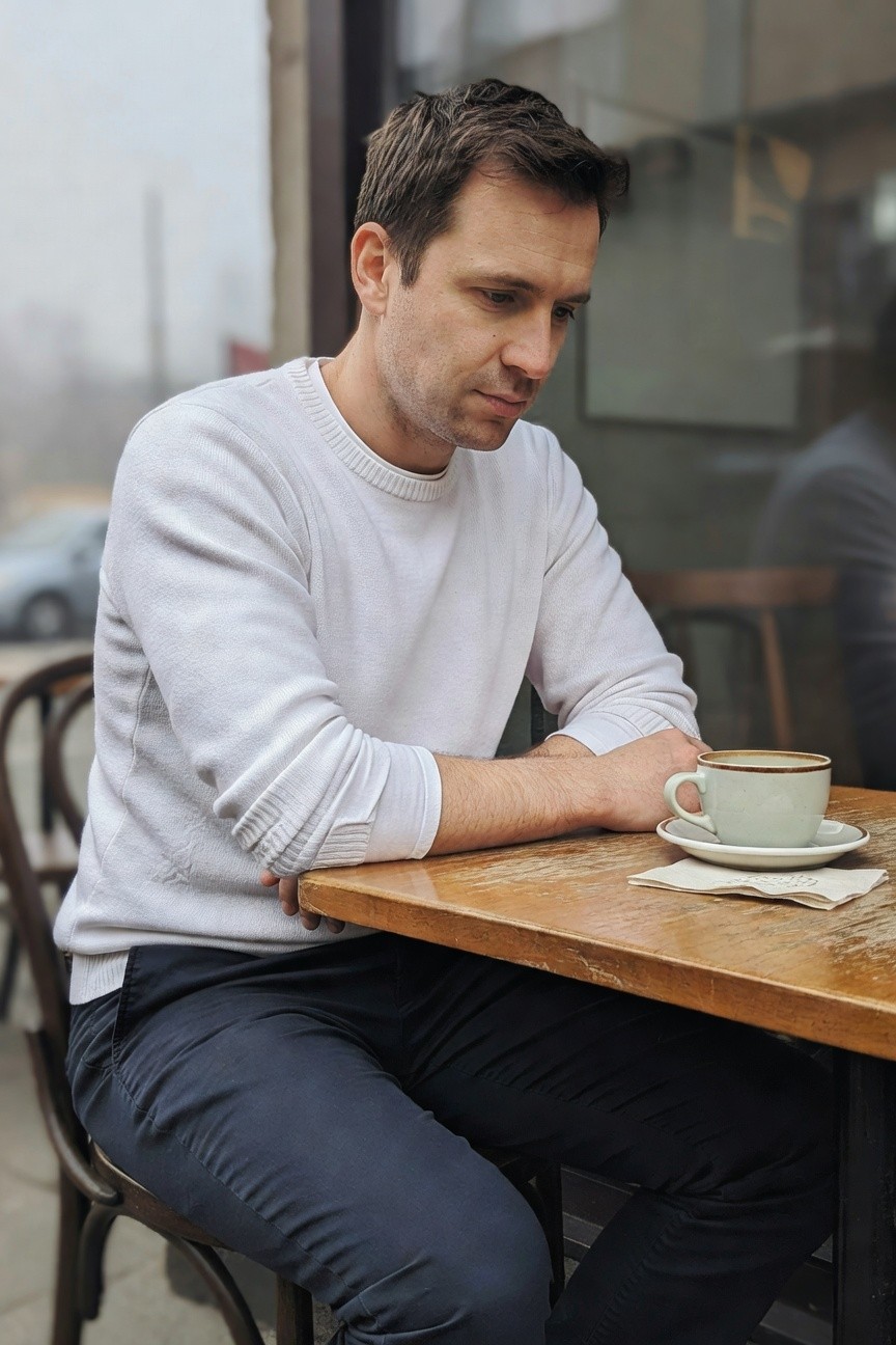 Man in a textured white crewneck sweater and slim navy pants sits at a wooden cafe table with arms crossed, holding a saucer and coffee cup nearby