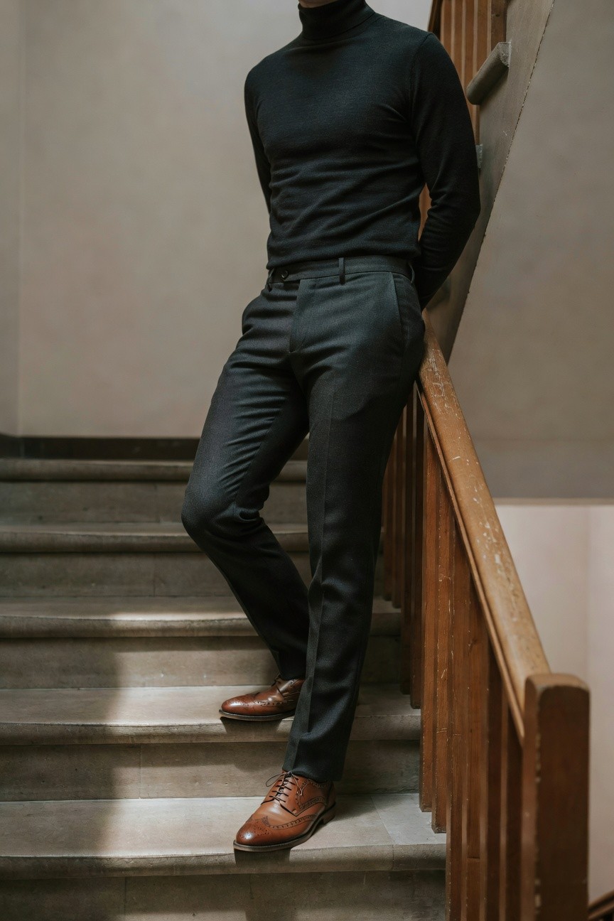 Man leaning casually on wooden staircase railing wearing black turtleneck sweater, slim charcoal grey trousers, and tan brogue leather shoes, indoor setting with stone steps.