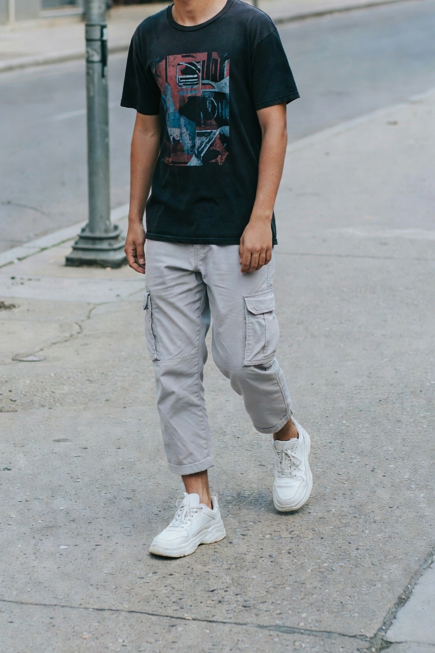 Young man in faded black graphic t-shirt with abstract red and blue design, light grey cargo pants cuffed at ankles, and white chunky sneakers, walking on urban sidewalk