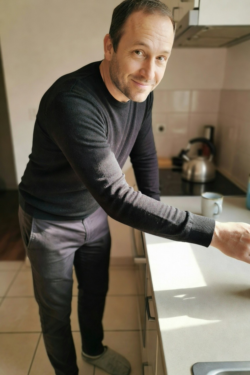 Man in black knit sweater and light grey slim pants leaning on kitchen counter holding a mug, wearing grey slippers, casual home style