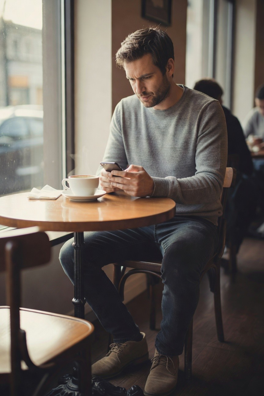 Man in light grey crewneck sweater, dark slim jeans, and tan suede lace-up boots, sitting at a wooden cafe table holding a phone, with white coffee cup nearby