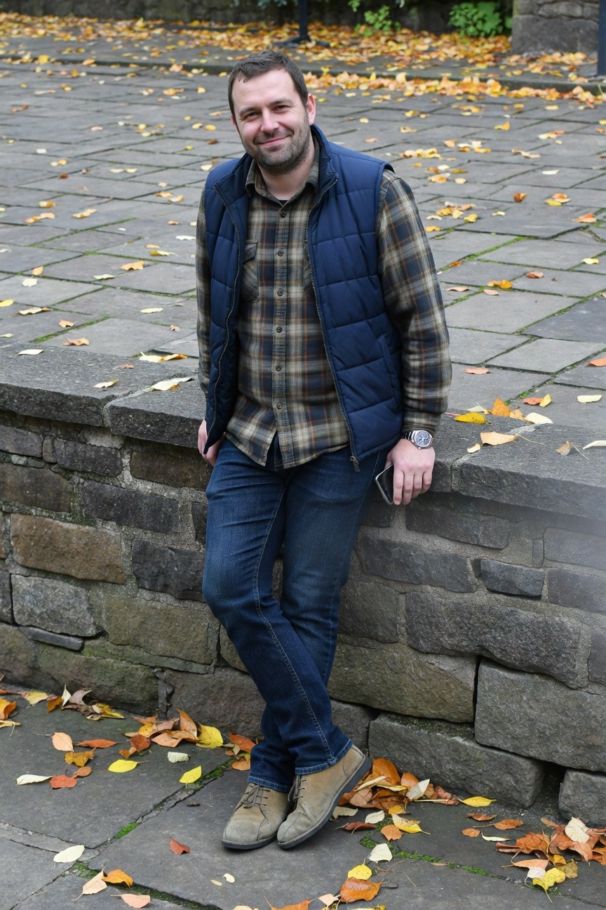 Man leaning against stone wall in layered plaid flannel shirt under navy puffer vest, dark slim jeans, tan suede boots, holding phone, autumn leaves scattered on pavement