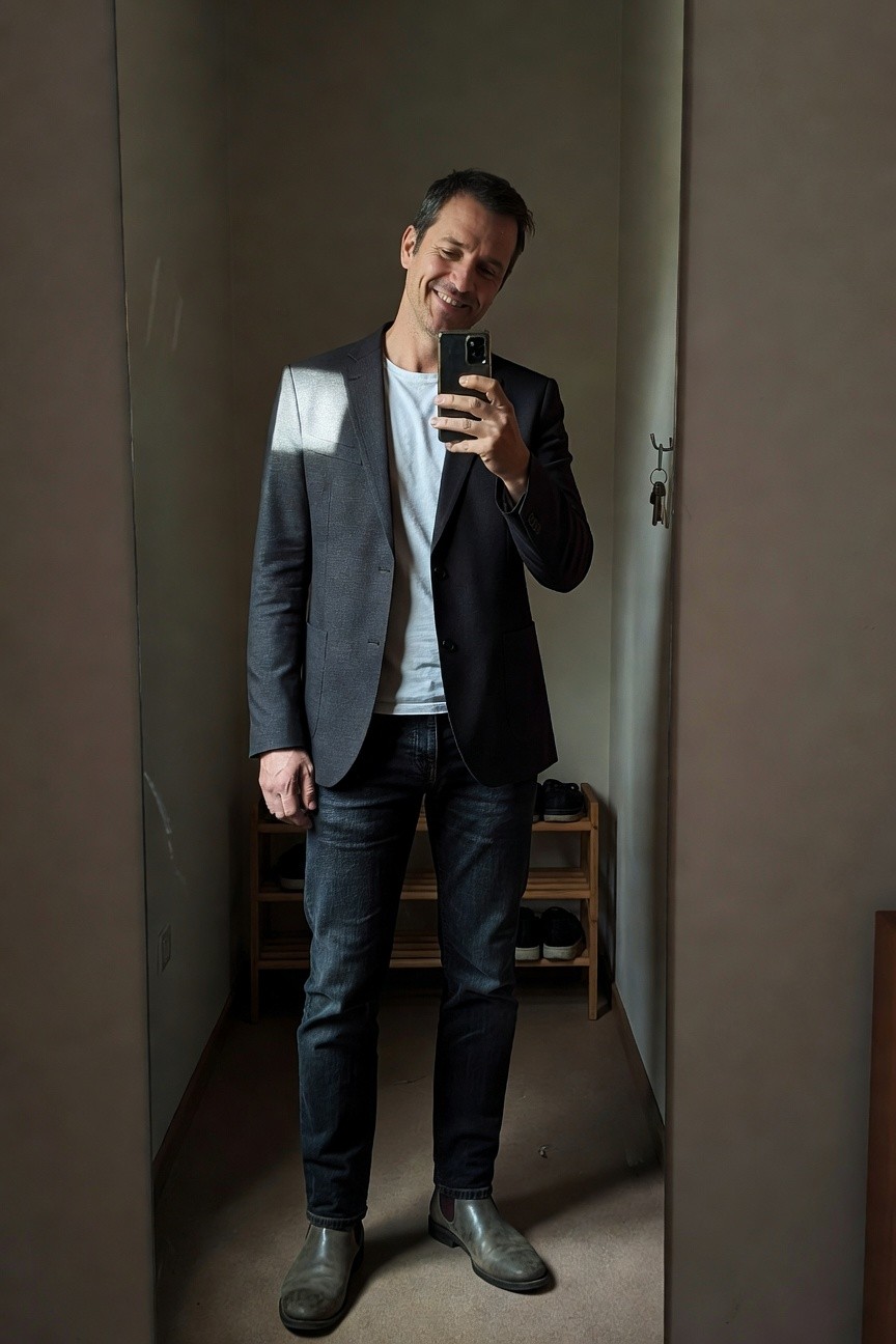 Man in a textured charcoal gray blazer over white t-shirt, slim dark blue jeans, and gray chelsea boots, standing in a closet mirror selfie smiling confidently