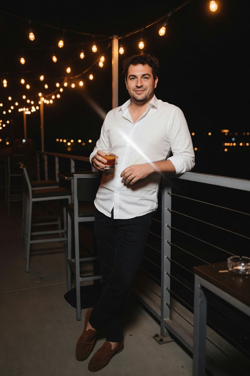 Man in white button-down shirt with rolled sleeves, slim dark jeans, and brown suede loafers, leaning on a railing while holding a glass of amber drink