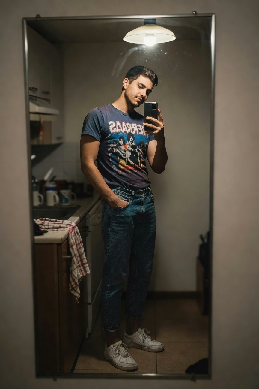 Mirror selfie of a man in a faded navy Motörhead band graphic t-shirt, medium blue relaxed straight-leg jeans, and white Nike Air Force 1 sneakers, hands in pockets, standing in a kitchen.