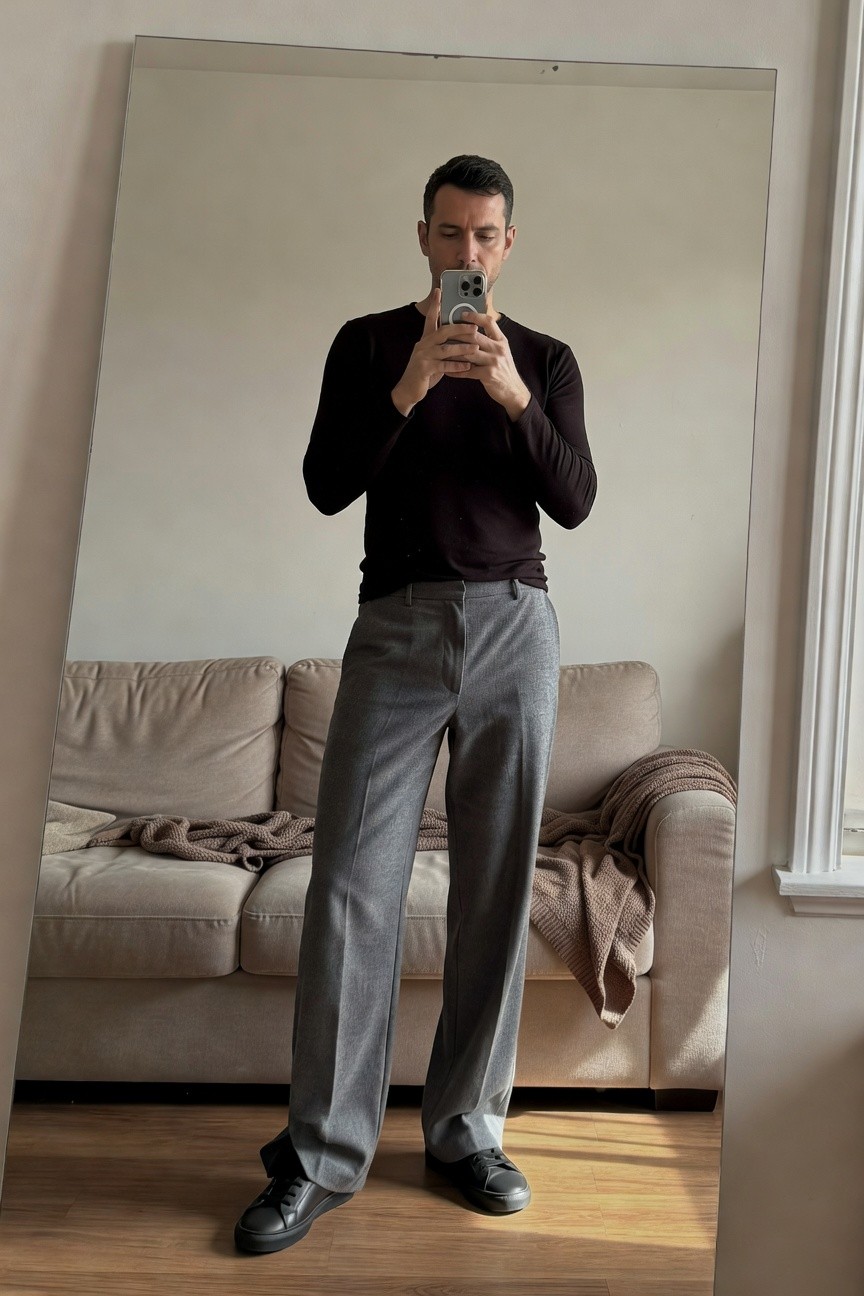 Man in fitted black long-sleeve sweater, wide-leg light grey trousers, and black sneakers, posing in front of a full-length mirror for a mirror selfie