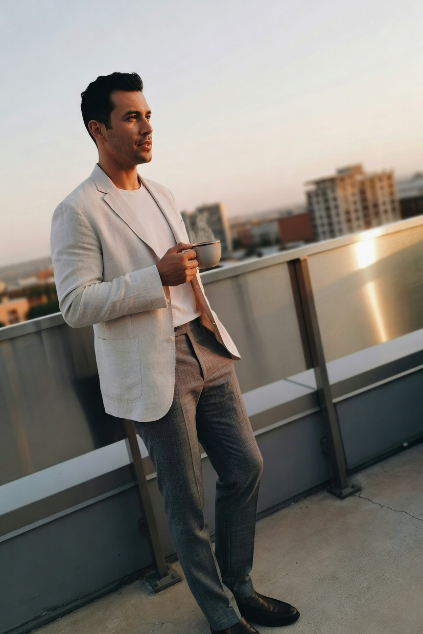 Man in cream linen blazer over white t-shirt, grey trousers, and black loafers, leaning against rooftop railing while holding a coffee mug
