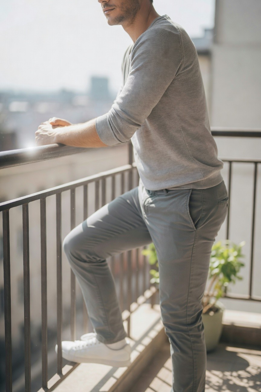 Side view of a man in a light grey long-sleeve sweater, matching darker grey slim pants, and white sneakers, leaning casually on a balcony railing.