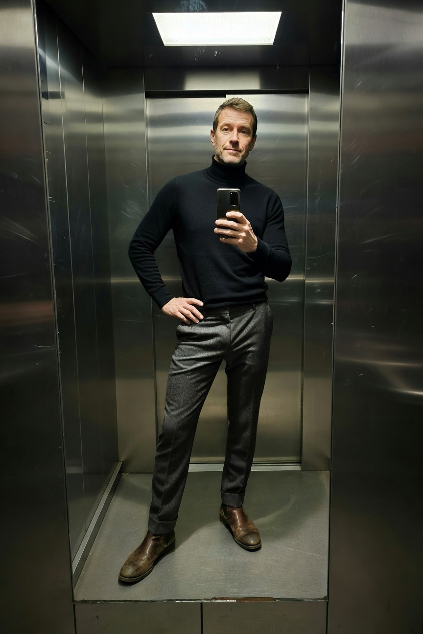 Man taking a mirror selfie in an elevator wearing a fitted black turtleneck sweater, slim grey wool trousers, and brown leather chelsea boots, standing confidently with one hand on hip