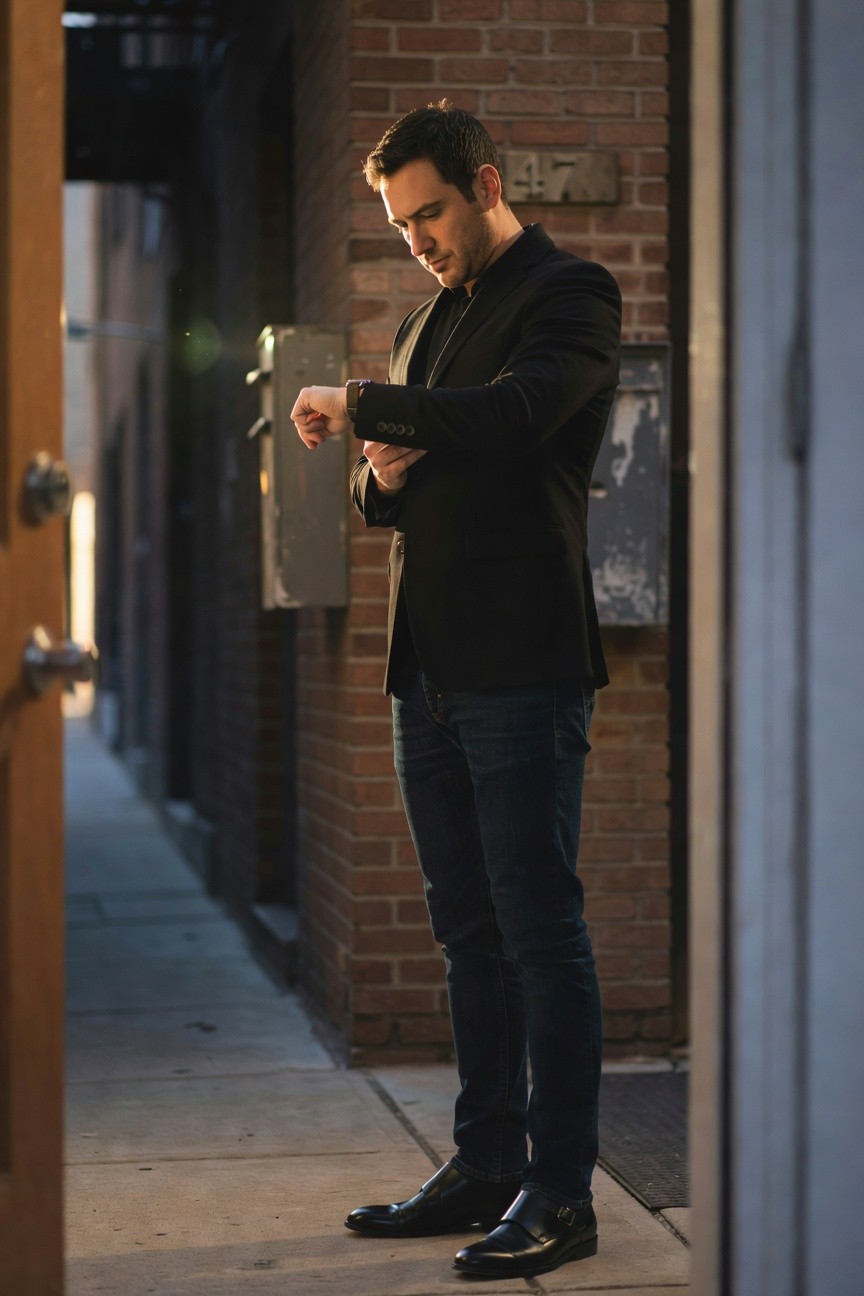 Side profile of a man in a slim black blazer, dark shirt, slim dark jeans, and black buckle dress shoes, adjusting his watch while standing near a door