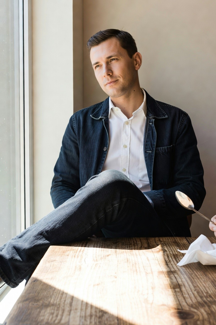 Man in dark denim jacket over white shirt and dark jeans, seated casually with one leg propped on a wooden table, holding a spoon and napkin