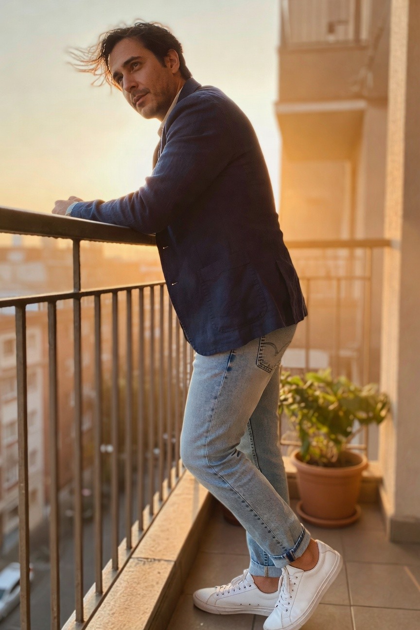 Man in navy unstructured blazer over white shirt, light wash slim jeans cuffed at ankles, and white sneakers, leaning on balcony railing with city view in background