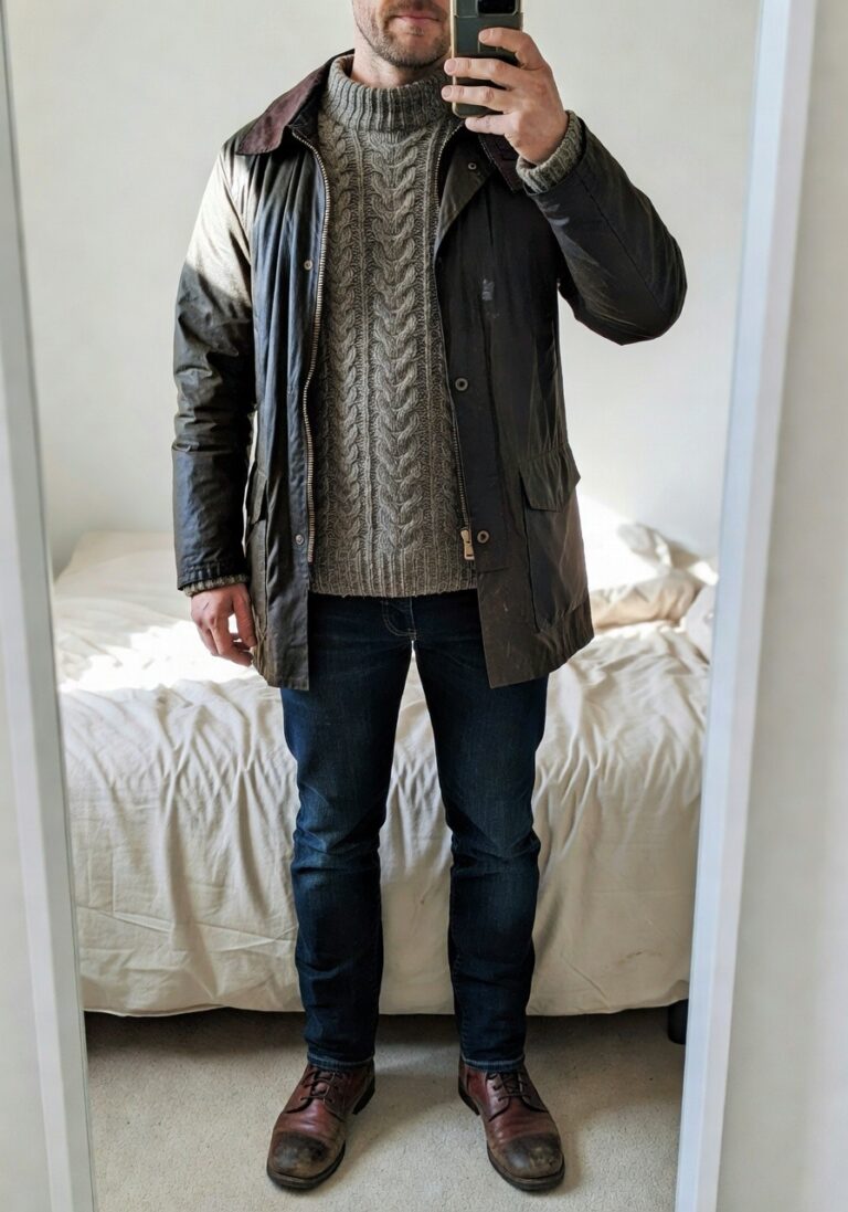 Mirror selfie of a man in olive green waxed cotton jacket layered over gray chunky cable knit turtleneck sweater dark slim jeans and reddish brown leather lace-up boots standing in a bedroom with white bed in background