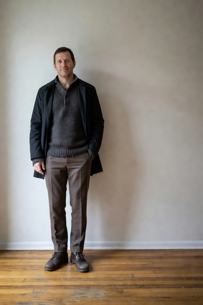 Man in navy overcoat layered over textured gray knit sweater, brown wool trousers, and rugged brown leather boots, standing casually with hands in pockets against plain white wall and wood floor