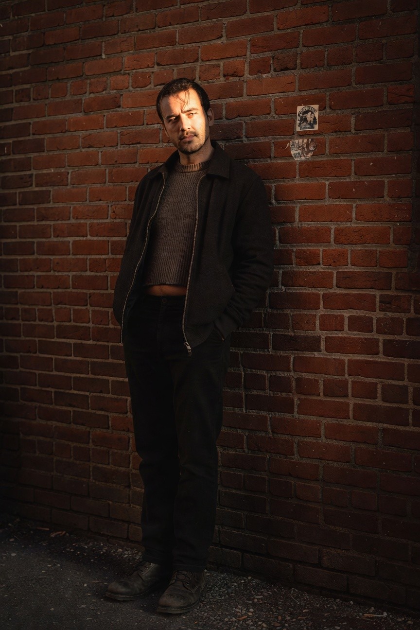 Man in open black quilted coat over gray cropped sweater, black slim pants, and black leather boots, leaning against brick wall with hands in pockets