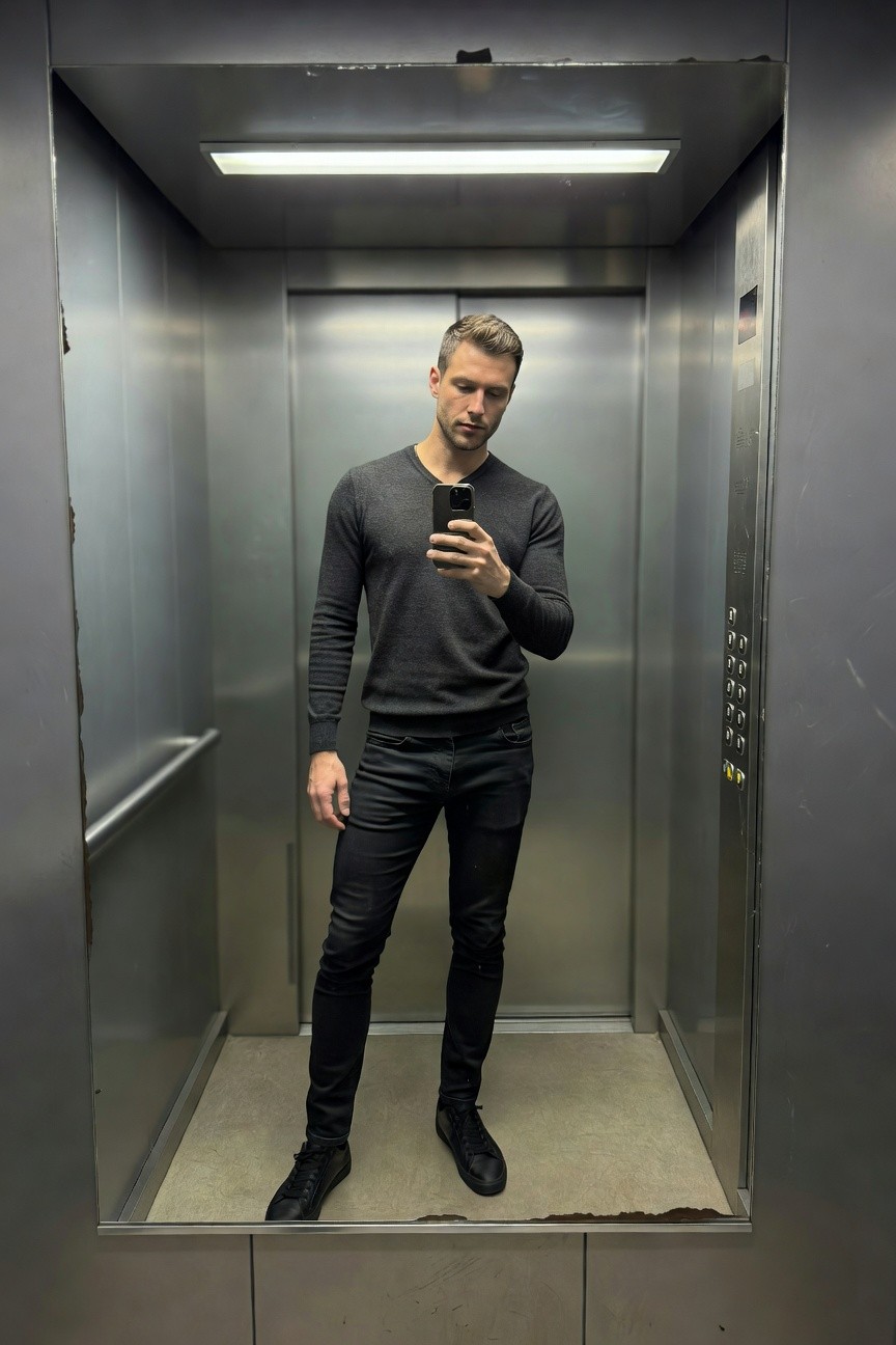 Man taking a mirror selfie in a steel elevator, wearing a gray V-neck long-sleeve sweater, black slim-fit jeans, and black sneakers, standing casually with one hand in pocket
