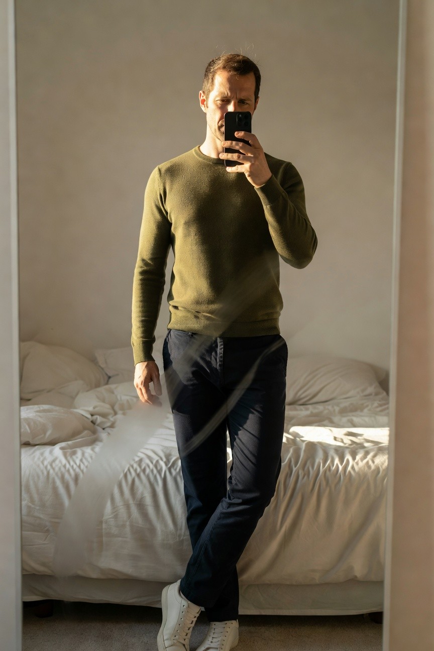 Man in mirror selfie wearing olive green crewneck sweater, navy slim trousers with drawstring, and white leather sneakers, standing casually in bedroom with bed visible
