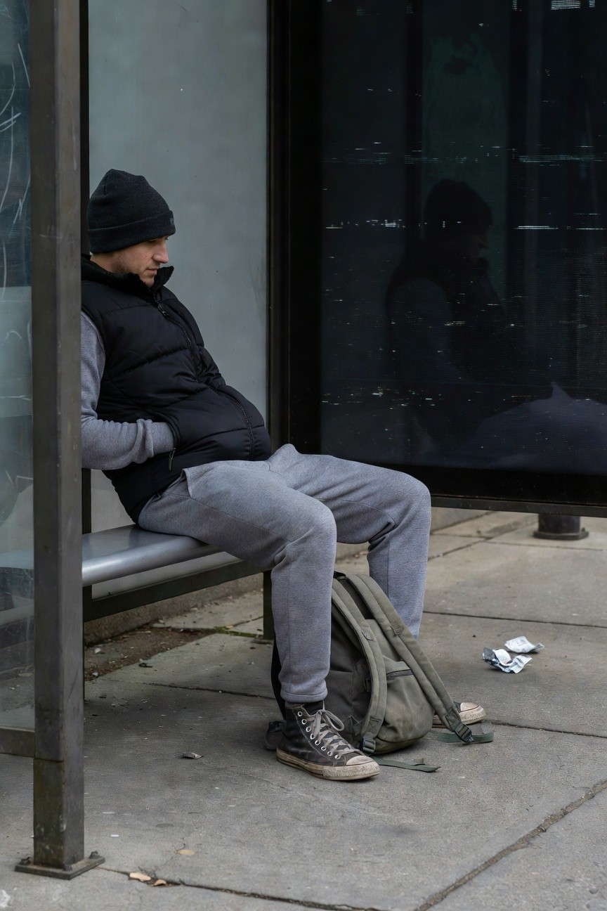 A man sits on a bus bench wearing grey joggers, black puffer vest over grey hoodie, black beanie, black Converse sneakers, and olive backpack nearby on grey pavement