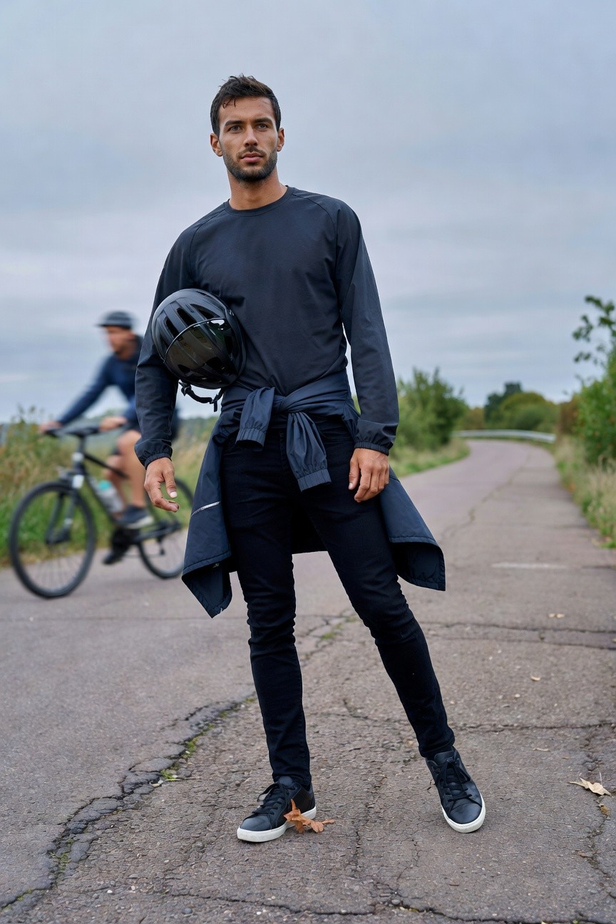 A man in slim black jeans, navy long-sleeve pullover, navy jacket tied around waist, black sneakers, holding black helmet, standing on cracked path with blurred cyclist behind