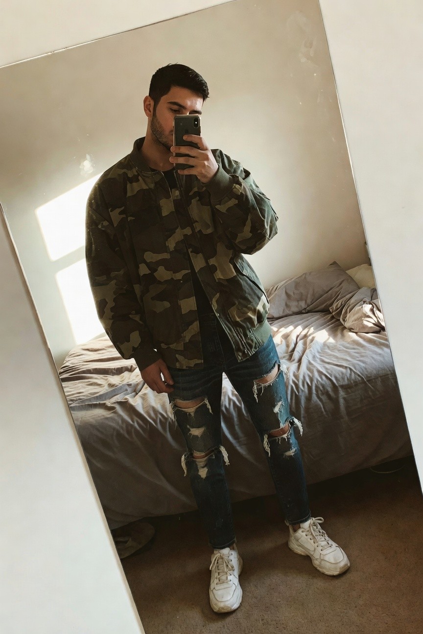 Man in green camo bomber jacket, black t-shirt, ripped blue jeans, and white sneakers posing in front of mirror selfie-style