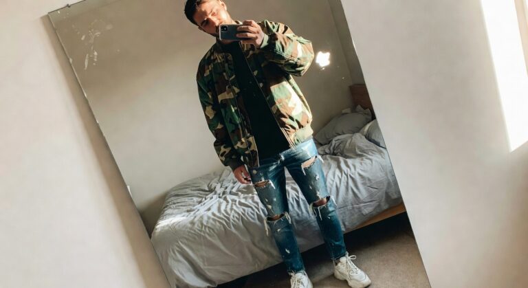 Man in green camo bomber jacket, black t-shirt, ripped blue jeans, and white sneakers posing in front of mirror selfie-style