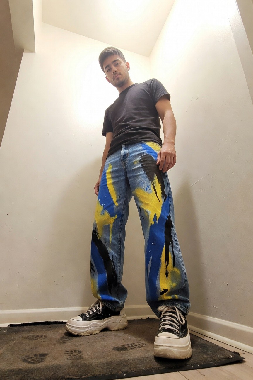 Young man in black short-sleeve t-shirt, blue jeans hand-painted with yellow and black abstract splatters, and white platform sneakers, posing confidently in a plain indoor corner