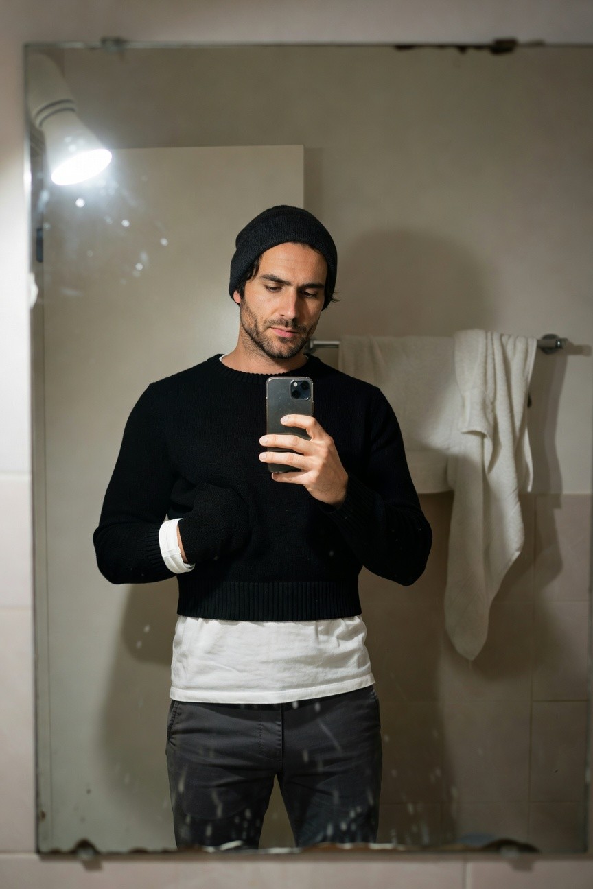 Mirror selfie of a man in a black beanie, black knit sweater layered over a white t-shirt with cropped hem, grey trousers, white-trimmed black wrist wraps, and black socks, standing in a tiled bathroom