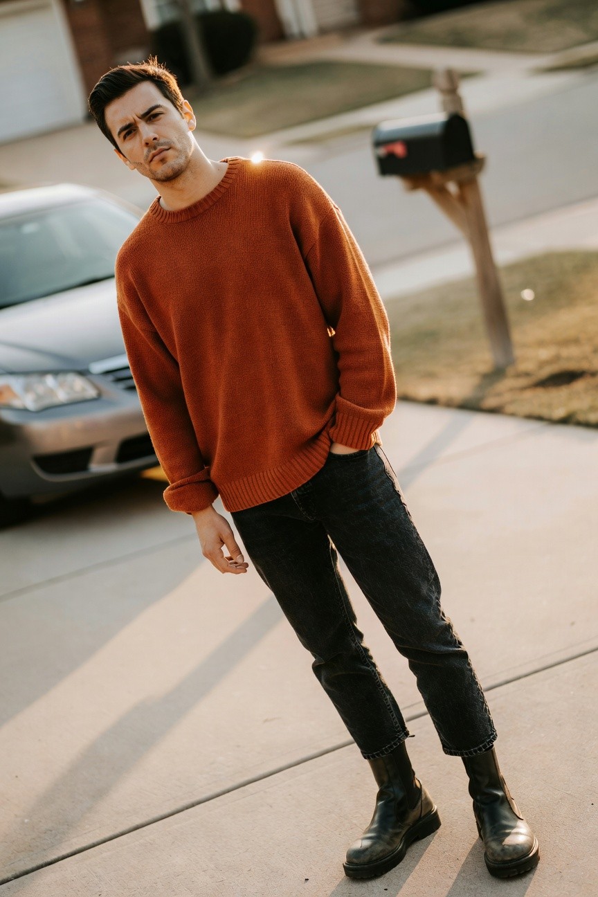 Man in oversized rust chunky knit crewneck sweater, slim black ankle-length jeans, and black platform Chelsea boots, posed casually for modern streetwear look