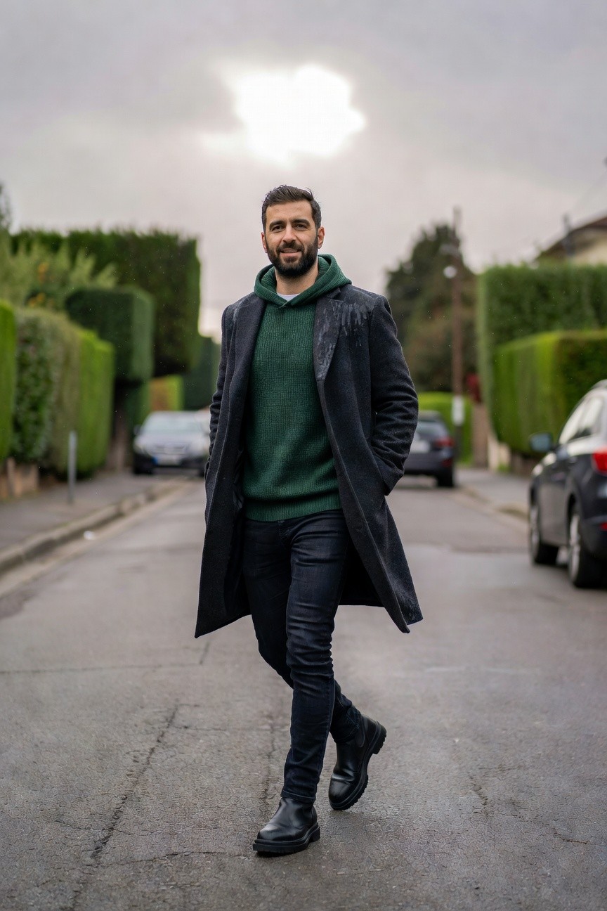 Man in green hoodie layered under long black wool overcoat, black slim jeans, and black Chelsea boots, walking on urban street