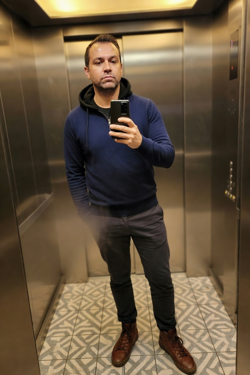 Man taking elevator selfie in navy hooded knit sweater, grey straight-leg trousers, and brown laced leather boots, hands casually posed