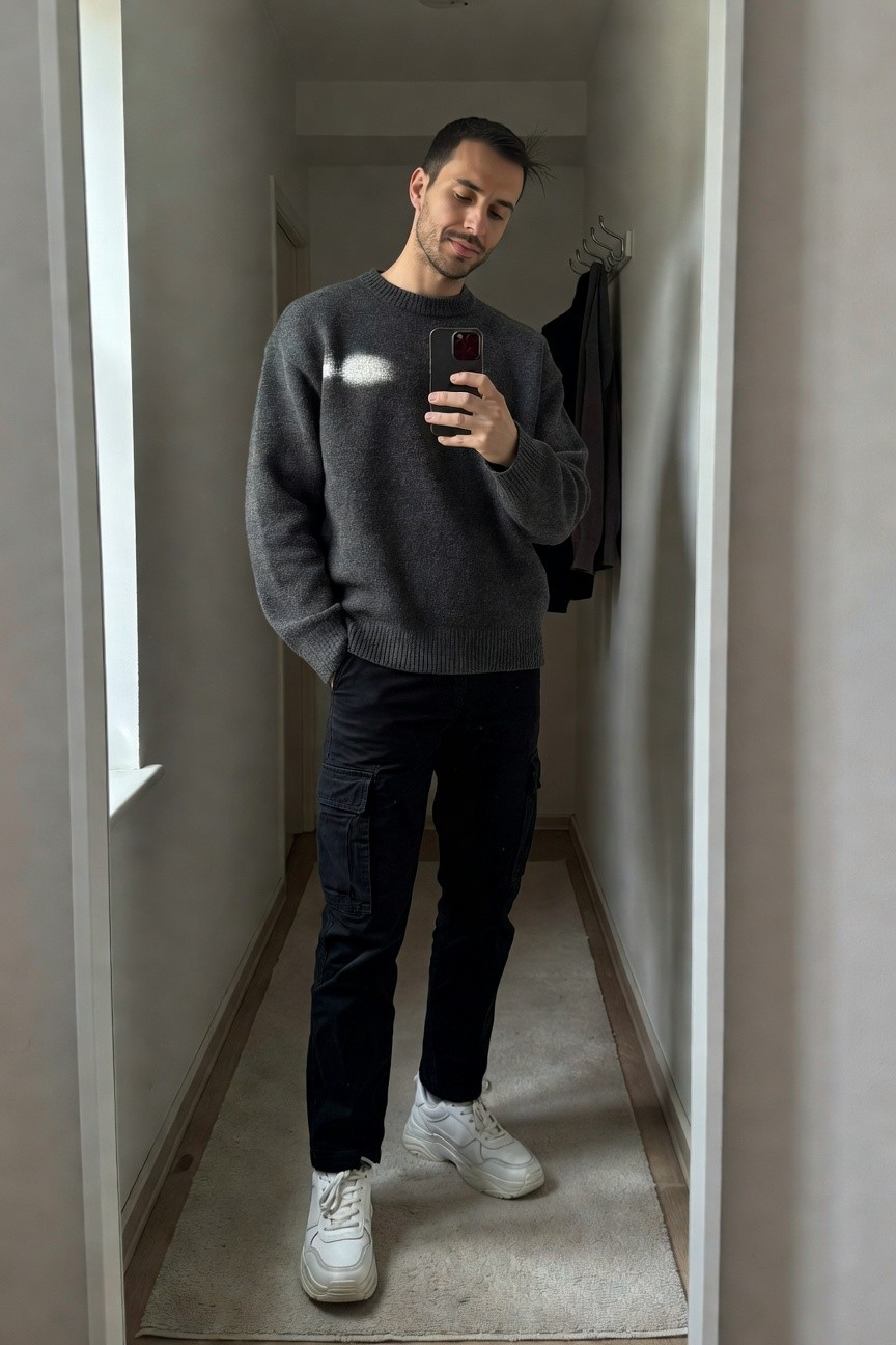Man taking mirror selfie in gray oversized knit sweater with subtle white mark, black straight-leg cargo pants, and chunky white sneakers, hands in pockets, hallway setting