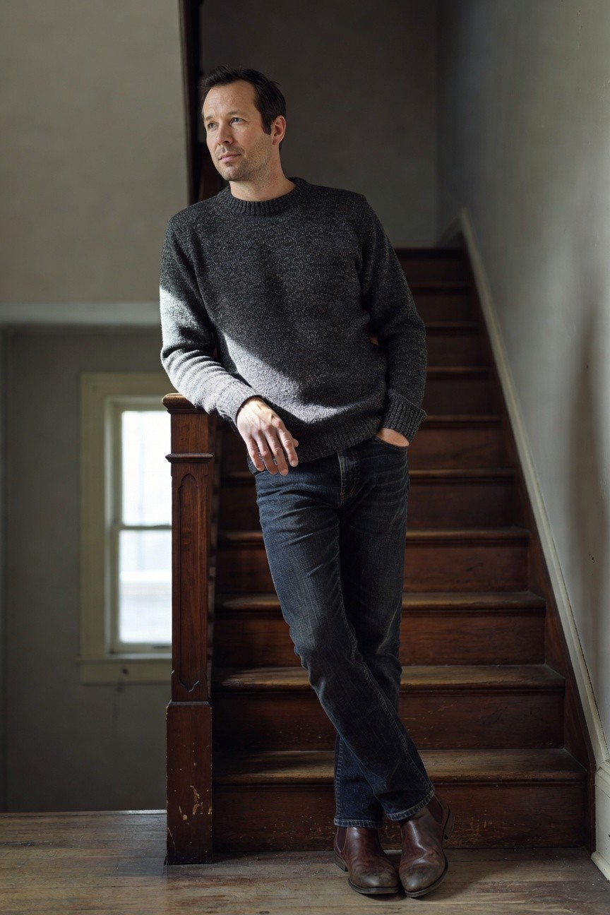 Man in textured gray crewneck sweater, dark straight-leg jeans, and brown leather chelsea boots, leaning casually on wooden railing with hands in pockets