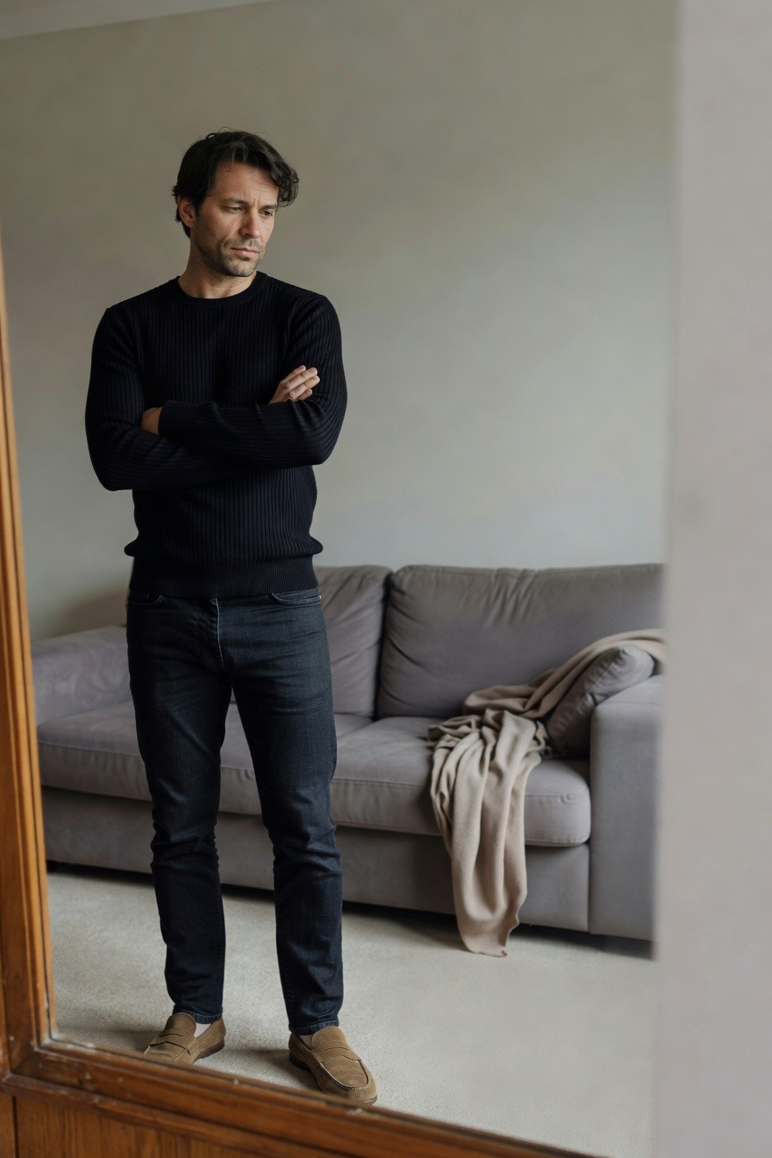 Man standing arms crossed in front of mirror wearing black ribbed crewneck sweater, slim dark jeans, and tan suede loafers in a minimalist indoor setting
