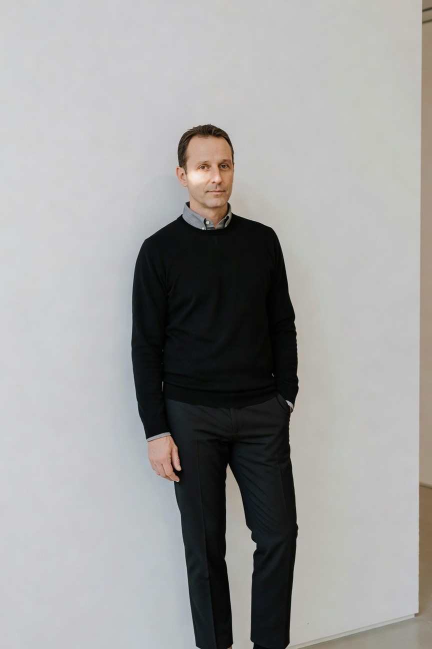 Man in black crewneck sweater layered over white collared shirt with slim black trousers, standing casually against plain wall, sharp minimal style