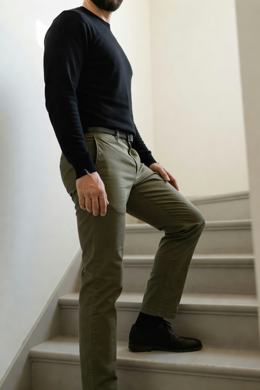 Side view of a man in a black long-sleeve crewneck sweater, olive green slim chinos, black leather belt, black socks, and black leather boots, standing on a staircase with hand in pocket