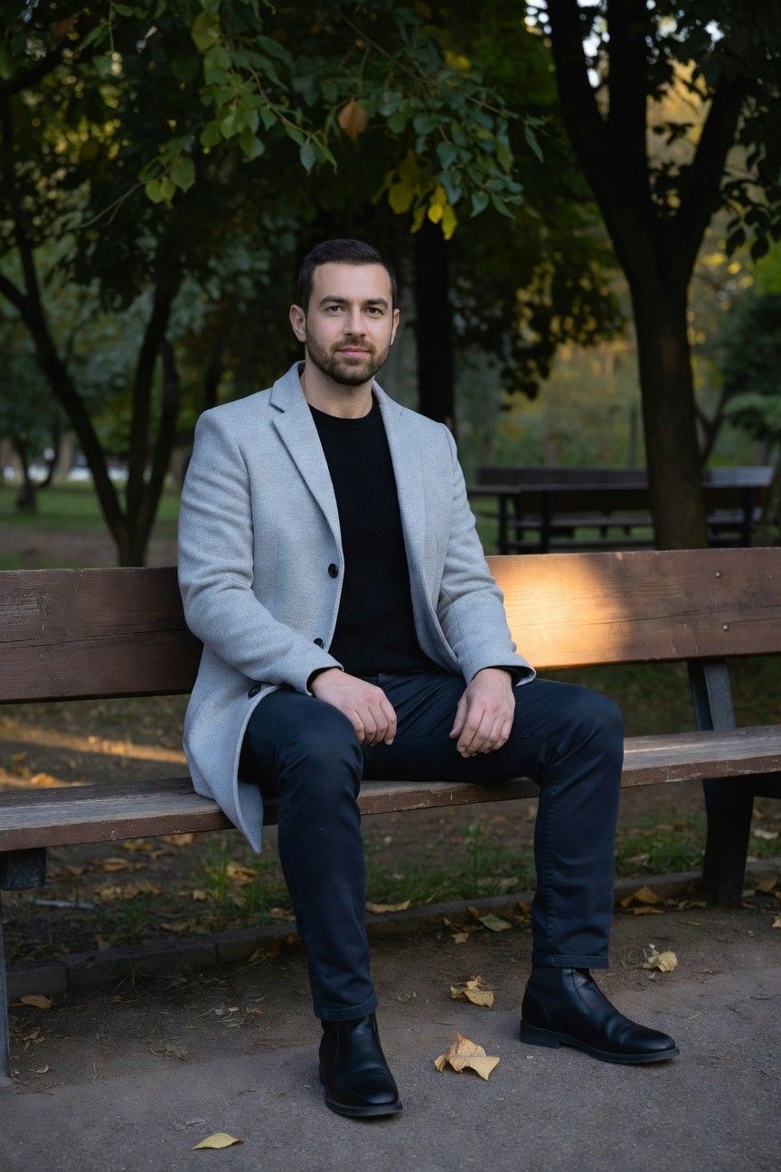 Man in light gray wool overcoat, black turtleneck sweater, slim black pants, and black leather ankle boots, sitting on a wooden park bench with hands clasped