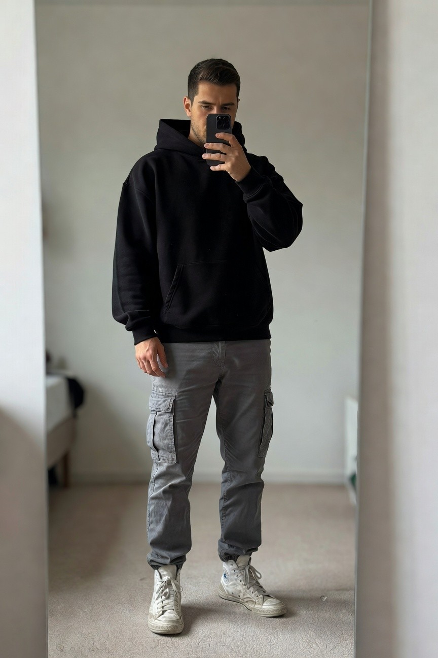 Mirror selfie of a man in an oversized black hoodie, grey cargo pants with multiple pockets, and white high-top sneakers, standing casually in a bedroom